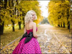 Blondynka w różowej sukni z czarną kokardą spacerująca parkową aleją