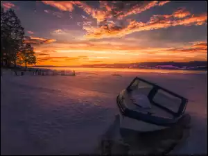 Motorówka nad ośnieżonym jeziorem z górami i drzewami w tle o zachodzie słońca