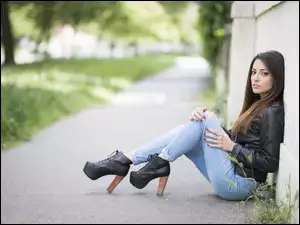Dziewczyna w skórzanej kurtce i butach siedzi oparta o ścianę budynku
