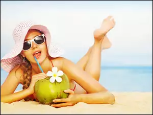 Kobieta w kapeluszu i okularach przeciwsłonecznych pije sok z owoca na plaży