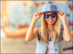 Uśmiechnięta dziewczyna o blond włosach w czapce i okularach przeciwsłonecznych