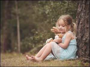 Dziewczynka tuląc pluszowego królika siedzi pod drzewem w lesie