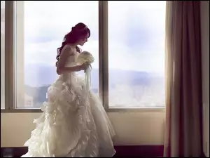 Panna młoda w białej sukni z bukietem w dłoniach przy stoi oknie