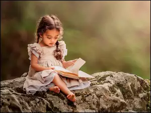 Dziewczynka z warkoczykami siedzi na kamieniu przeglądając książkę