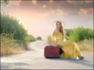 Kobieta w kapeluszu i letniej sukience na drodze z walizką