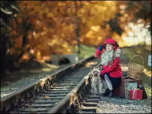 Dziewczynka z pieskiem siedzi na walizkach przy torach kolejowych