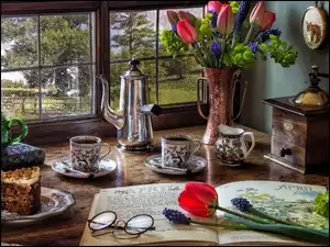 Kwiaty w wazonie książka i filiżanki postawione obok lampy i dzbanka na stole