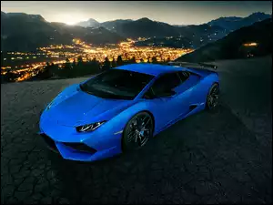 Niebieski samochód marki Lamborghini na tle oświetlonego miasta o zmroku u stóp gór