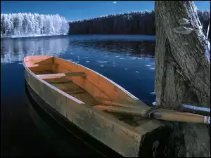 Łódka przypięta do drzewa na tle ośnieżonych lasów nad jeziorem
