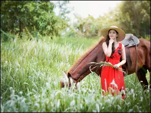 Kobieta w kapeluszu obok konia pasącego się w trawie