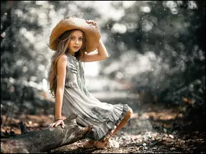 Dziewczynka w plecionym kapeluszu siedzi na kłodzie