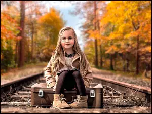 Dziewczynka siedzi na walizce w środku torów kolejowych