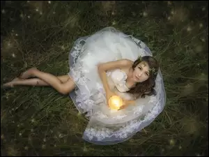 Kobieta wbiałej sukience ze święcą w rękach leży na trawie