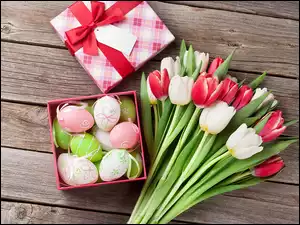 Pisanki w pudełku obok białych i czerwonych tulipanów na deskach