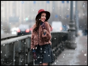 Uśmiechnięta modelka w kapeluszu na moście obserwuje prószący śnieg