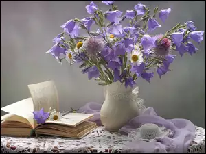 Kompozycja z książki obok wazonu z dzwonkami i margerytkami