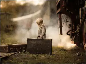 chłopiec przy torach z lokomotywą i trzymający starą walizkę