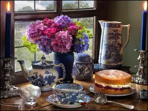 Bukiet hortensji przy oknie obok świec, ciasta i porcelanowych naczyń