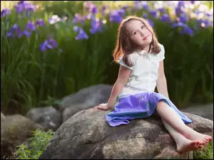 Dziewczynka siedzi na kamieniu z kwiatami irysów w tle