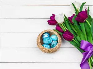 Niebieskie pisanki w koszyczku obok bukietu tulipanów z wstążką