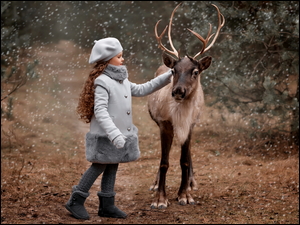 Dziewczynka w berecie i szarym płaszczyku głaszcząca renifera w lesie