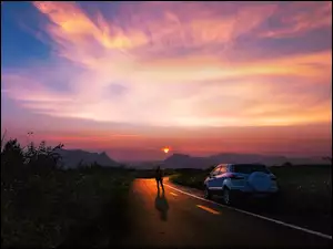 Człowiek obok samochodu na drodze i zachód słońca w tle