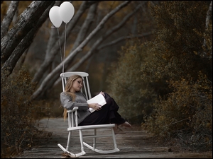 Dziewczyna czytająca książkę siedząc w fotelu na alejce między drzewami