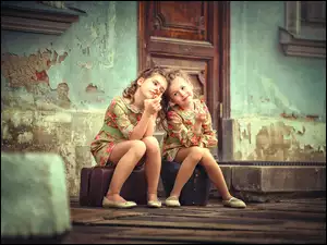 Dwie dziewczynki siedzą na torbach przed zniszczonym domem