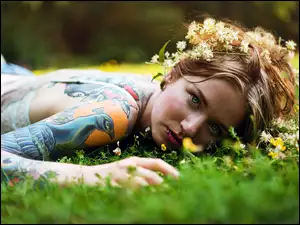 Blondynka z tatuażami i wiankiem na głowie leżąca na trawie