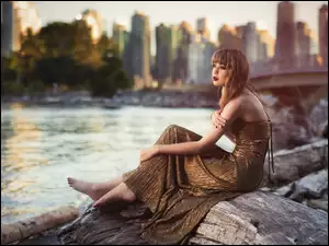 Kobieta w długiej sukience boso siedzi na kłodzie na brzegu rzeki