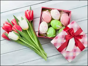 Białe i czerwone tulipany obok pudełka z pisankami na jasnych deskach