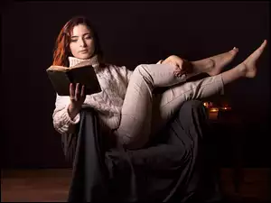 Kobieta siedząca w fotelu z książką w ręku na ciemnym tle