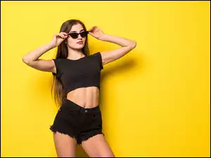 Dziewczyna w czarnym stroju i w okularach przeciwsłonecznych na żółtym tle
