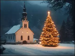 Rozświetlona światełkami choinka na śniegu przed kościołem na tle gór