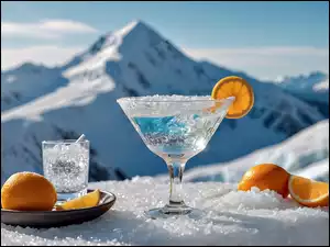 Na śniegu na tle gór pucharek z lodem i pokrojonym plasterkiem pomarańczy do picia