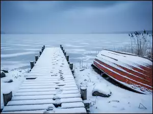 Odwrócona łódka przyośnieżonym pomoście nad zamarzniętym jeziorem pod zachmurzonym niebem