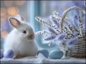 Biały królik i niebieskie pisanki obok koszyczka z baziami i kłosami